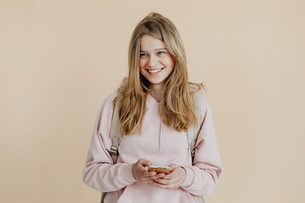 A cheerful teenager with blonde hair wearing a pink sweater and using a smartphone indoors.